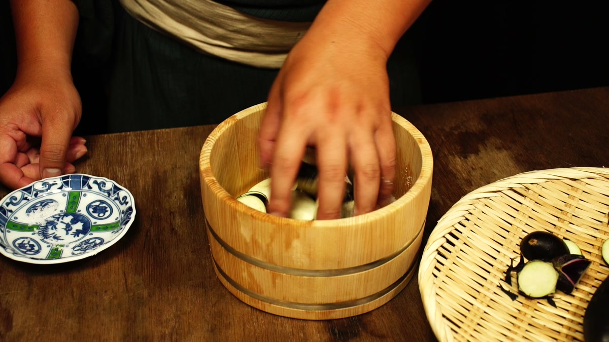 木桶に入れた輪切りの茄子を手でもみこんでいるところ。傍らに竹ざるの茄子