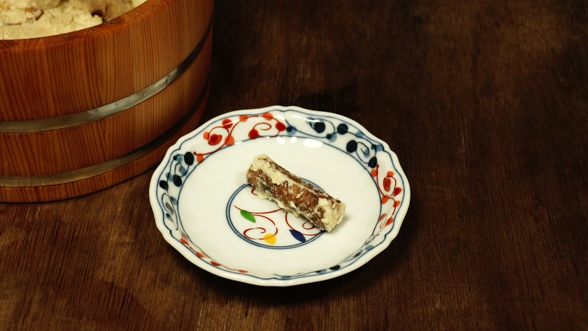 色絵の小皿にのせた牛蒡の粕漬け一切れ。奥に木桶が見える