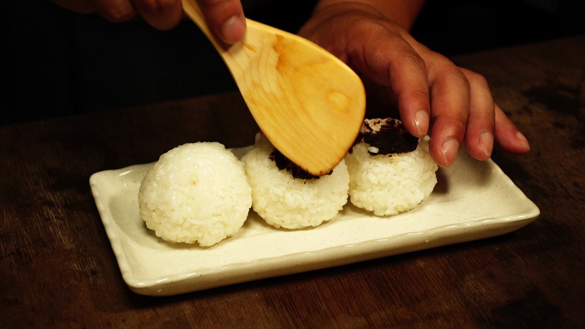 皿に並べた三つの握り飯に、しゃもじで豆味噌を塗っているところ