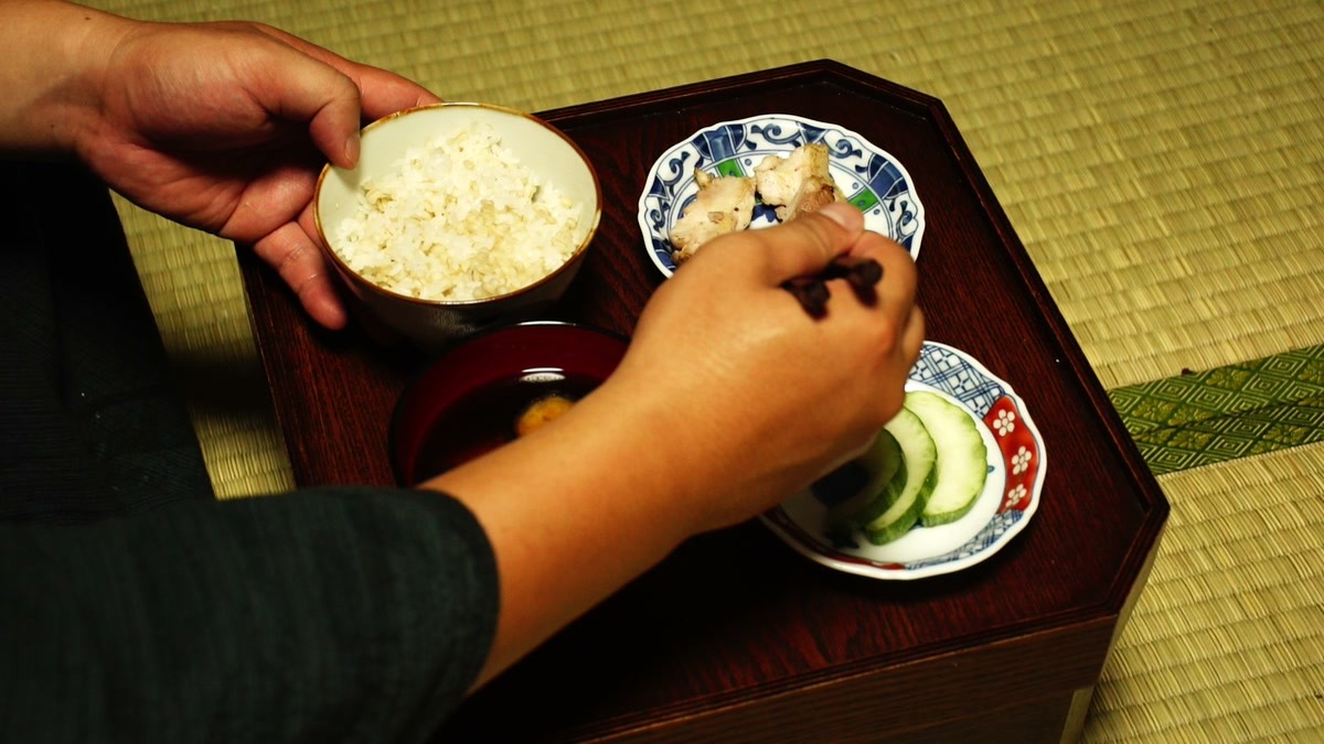 畳の上のお膳から麦飯の椀を手に取り、実際に食べているところ